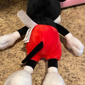 Puede incluir: Un peluche de Mickey Mouse, visto desde atr&aacute;s. El juguete presenta una cabeza y orejas negras, guantes y pies blancos y pantalones cortos rojos. Una cola negra se extiende desde la parte posterior. Una etiqueta blanca est&aacute; adherida al juguete.