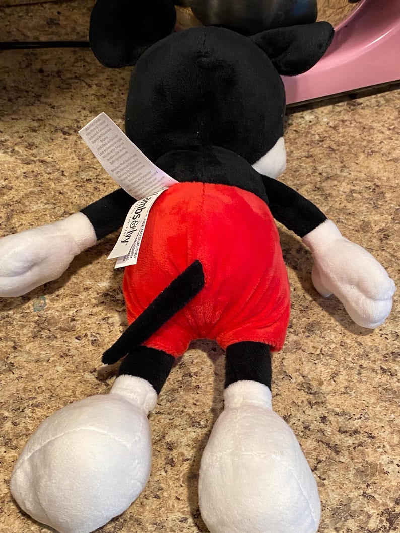 Puede incluir: Un peluche de Mickey Mouse, visto desde atr&aacute;s. El juguete presenta una cabeza y orejas negras, guantes y pies blancos y pantalones cortos rojos. Una cola negra se extiende desde la parte posterior. Una etiqueta blanca est&aacute; adherida al juguete.