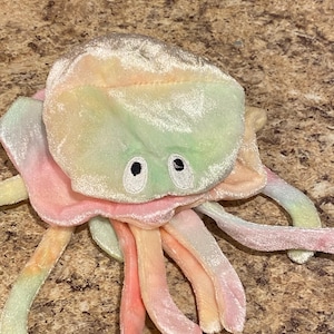 May include: A plush rainbow-colored jellyfish toy with embroidered eyes. The jellyfish has a soft, velvety texture and is a bright, cheerful color.
