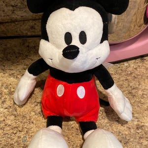 Puede incluir: Un peluche de Mickey Mouse. El juguete tiene una cabeza negra con orejas grandes, una cara blanca y una nariz negra. Lleva pantalones cortos rojos con botones blancos, brazos y piernas negros y guantes y zapatos blancos. El juguete es suave.