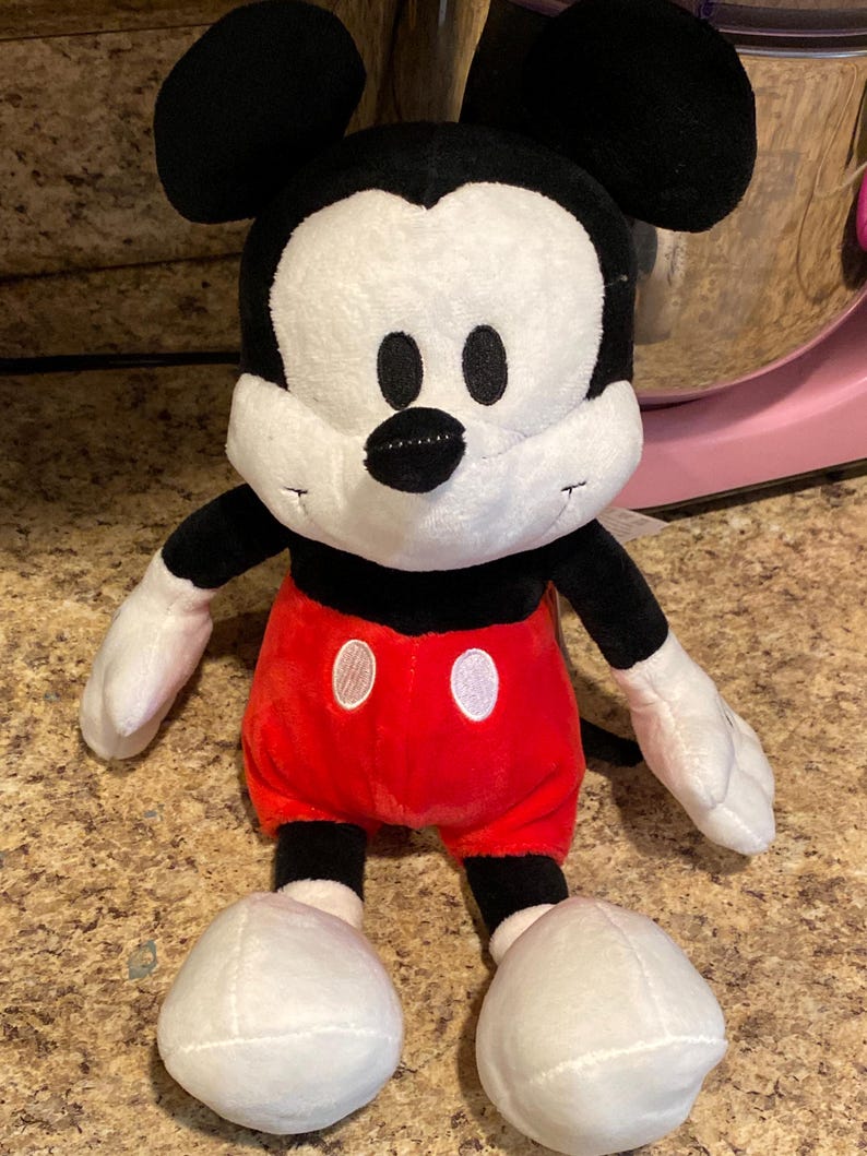 Puede incluir: Un peluche de Mickey Mouse. El juguete tiene una cabeza negra con orejas grandes, una cara blanca y una nariz negra. Lleva pantalones cortos rojos con botones blancos, brazos y piernas negros y guantes y zapatos blancos. El juguete es suave.