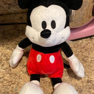 Puede incluir: Un peluche de Mickey Mouse. El juguete tiene una cara blanca, orejas negras y un cuerpo negro. Viste pantalones cortos rojos con botones blancos, guantes blancos y zapatos blancos. El dise&ntilde;o est&aacute; basado en el famoso personaje de Disney.