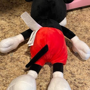 Puede incluir: Un peluche de Mickey Mouse, visto desde atr&aacute;s, con cabeza y orejas negras, guantes y pies blancos, y pantalones cortos rojos. El juguete tiene una cola negra y una etiqueta adjunta en la parte posterior.
