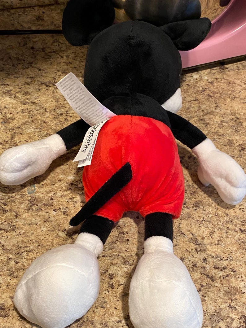 Puede incluir: Un peluche de Mickey Mouse, visto desde atr&aacute;s, con cabeza y orejas negras, guantes y pies blancos, y pantalones cortos rojos. El juguete tiene una cola negra y una etiqueta adjunta en la parte posterior.