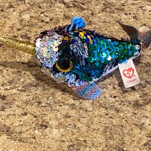 May include: A blue and green plush narwhal with iridescent sequins. The narwhal has a gold horn and large, glittery eyes. The plush toy has a Ty tag that reads "Calypso".