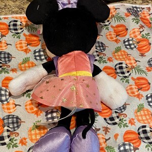 以下が含まれることがあります： ミニーマウスのぬいぐるみ。後ろから見て、黒い耳、紫のリボン、金色の星のアクセントが付いたピンクのドレスを着ています。人形は白い手袋と紫色の靴を履いています。背景はカボチャ模様の生地です。