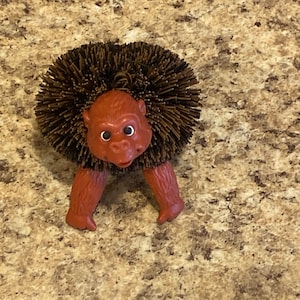 May include: A brown and red toy monkey with a brown bristly hair-like texture on its head. The monkey has a wide mouth and large eyes.