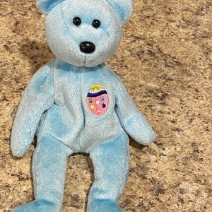 May include: A light blue beanie baby bear with a black nose and black eyes. The bear is wearing a pink and blue Easter egg patch on its chest.