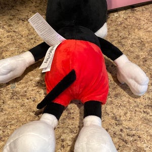 Puede incluir: Un peluche de Mickey Mouse, visto desde atr&aacute;s. El juguete presenta una cabeza y orejas negras, guantes blancos y pantalones cortos rojos. La cola es negra y los pies son grandes y blancos. Una etiqueta blanca est&aacute; adjunta.
