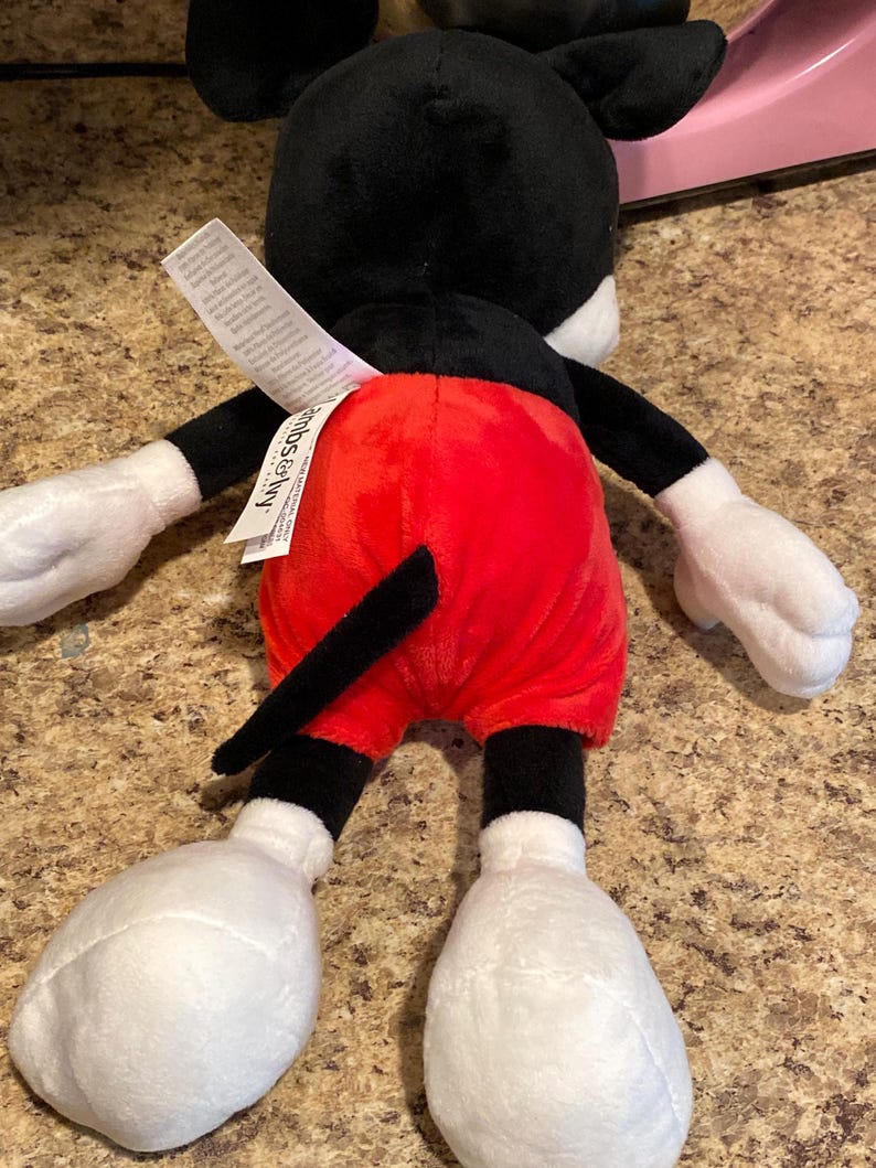 Puede incluir: Un peluche de Mickey Mouse, visto desde atr&aacute;s. El juguete presenta una cabeza y orejas negras, guantes blancos y pantalones cortos rojos. La cola es negra y los pies son grandes y blancos. Una etiqueta blanca est&aacute; adjunta.