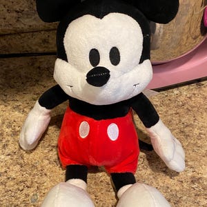 Puede incluir: Un peluche de Mickey Mouse. El personaje ic&oacute;nico tiene una cara blanca, orejas negras y un cuerpo negro. Lleva pantalones cortos rojos con detalles de botones blancos y guantes y zapatos blancos. El juguete es suave y tierno.