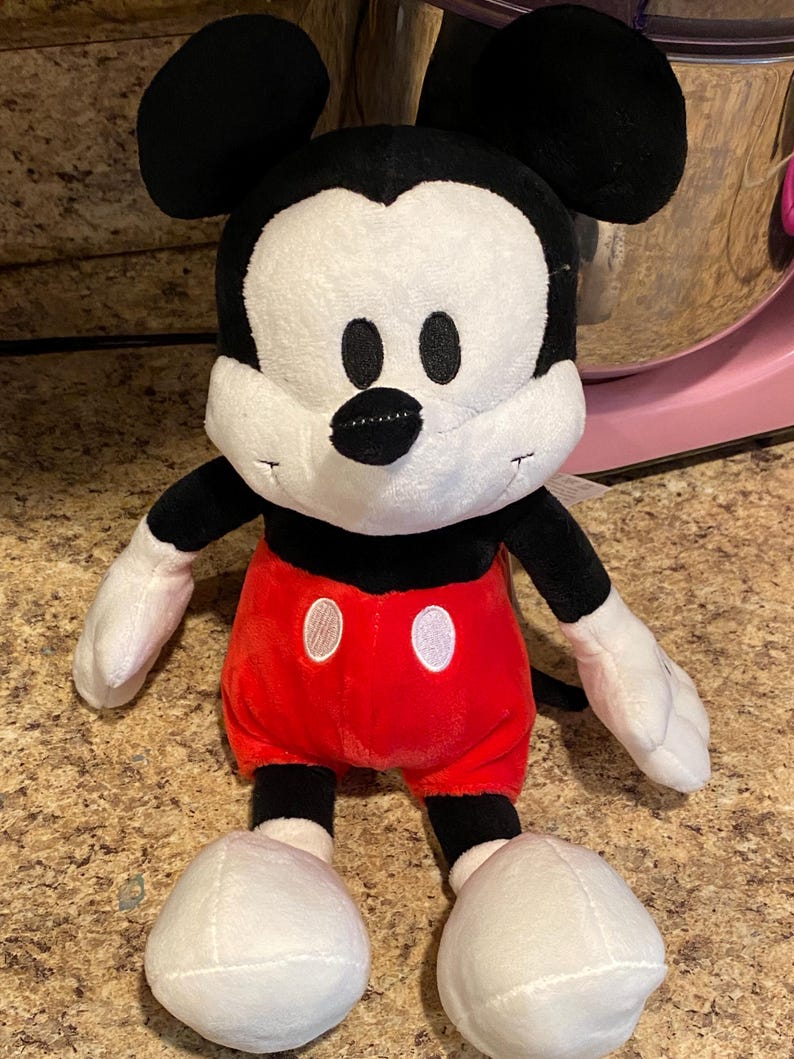 Puede incluir: Un peluche de Mickey Mouse. El personaje ic&oacute;nico tiene una cara blanca, orejas negras y un cuerpo negro. Lleva pantalones cortos rojos con detalles de botones blancos y guantes y zapatos blancos. El juguete es suave y tierno.