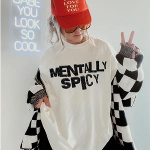 K&ouml;nnte beinhalten: Eine Person tr&auml;gt eine rote Trucker-M&uuml;tze mit dem Text "MAD LOVE FOR YOU" und einen wei&szlig;en Pullover mit dem Aufdruck "MENTALLY SPICY". Die Person tr&auml;gt auch eine schwarz-wei&szlig; karierte Strickjacke.