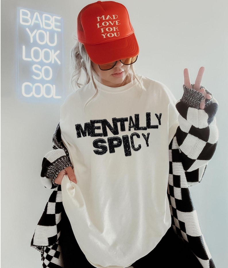 K&ouml;nnte beinhalten: Eine Person tr&auml;gt eine rote Trucker-M&uuml;tze mit dem Text "MAD LOVE FOR YOU" und einen wei&szlig;en Pullover mit dem Aufdruck "MENTALLY SPICY". Die Person tr&auml;gt auch eine schwarz-wei&szlig; karierte Strickjacke.