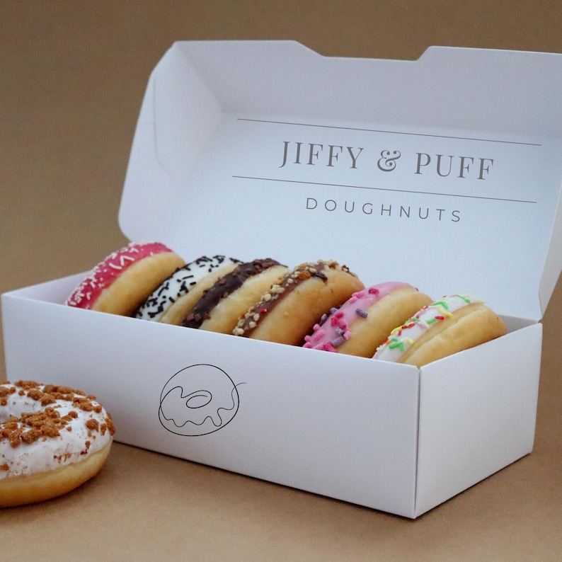 May include: A white box with the text "Jiffy & Puff Doughnuts" on the top. The box is open and contains six assorted donuts with different toppings. The donuts are arranged in a row, with the pink donut on the left and the white donut on the right.