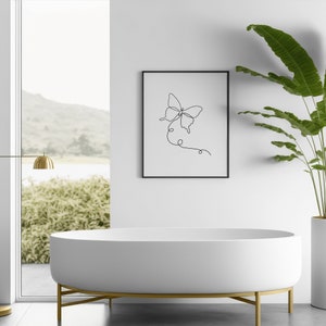 May include: A white bathtub with gold legs sits in a bathroom with a framed black and white line drawing of a butterfly on the wall. A potted plant is on the right side of the image.