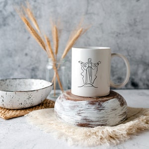 Puede incluir: Taza de cer&aacute;mica blanca con un dibujo de l&iacute;nea negra de una persona con los brazos extendidos. La taza est&aacute; sobre una base de madera blanca.