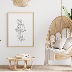 Puede incluir: Un dibujo en blanco y negro de una mujer con una t&uacute;nica larga, enmarcado en un marco de madera clara, colgado en una pared blanca. Una silla colgante de mimbre con una manta blanca est&aacute; en primer plano.