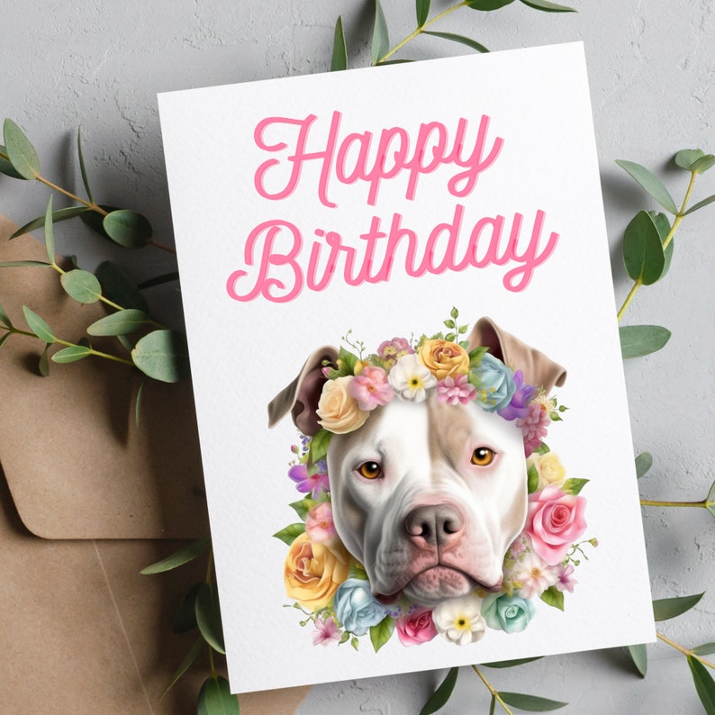 May include: Happy Birthday card featuring a white and brown pit bull wearing a flower crown. The flowers are pink, yellow, blue, and white.