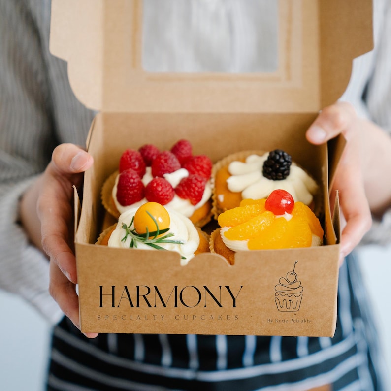 May include: A brown box with a clear window, containing four specialty cupcakes. The cupcakes are topped with raspberries, blueberries, cherries, and orange slices. The box has the text "HARMONY SPECIALTY CUPCAKES" and a logo of a cupcake with a cherry on top. The text "By Kyne Petraikis" is also on the box.