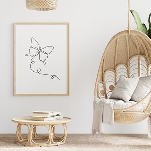 May include: A black and white line drawing of a butterfly on a white background in a wooden frame. The drawing is hanging on a white wall. A wicker hanging chair with a white cushion is in the foreground.