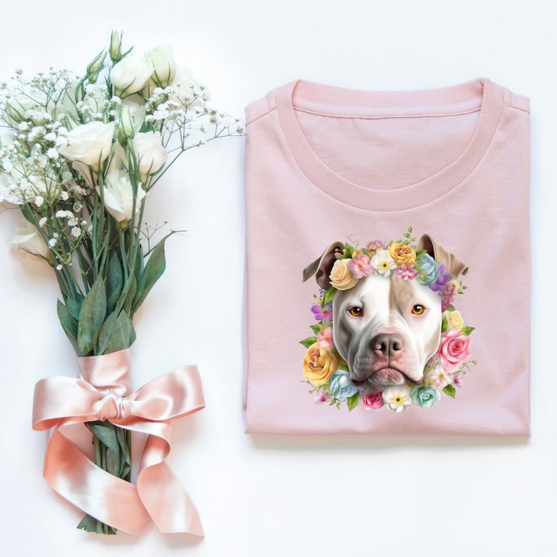 May include: A pink t-shirt with a white pit bull wearing a flower crown. The flowers are pink, yellow, blue, and purple.
