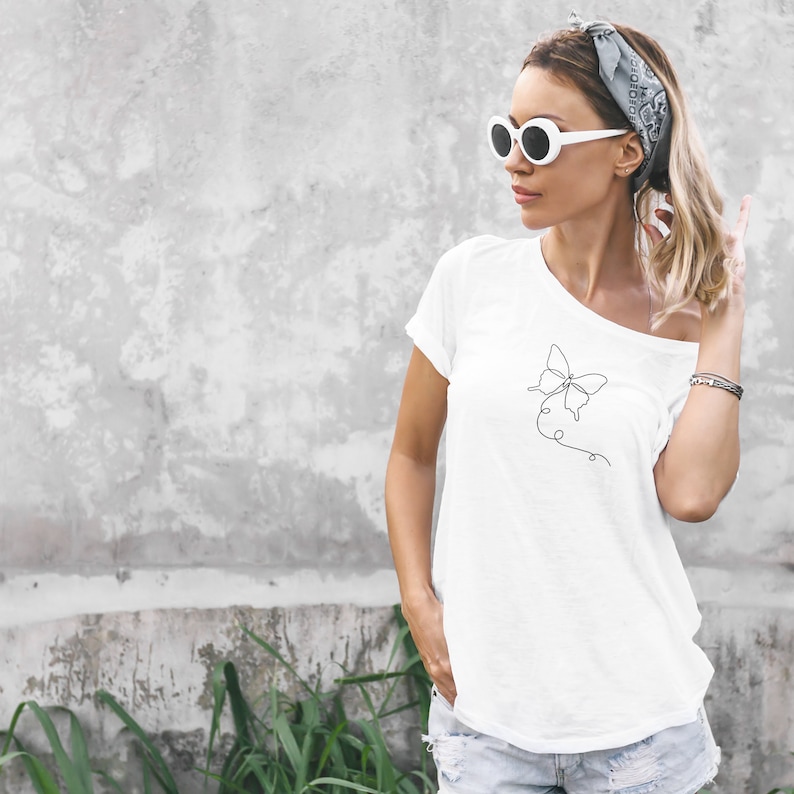 May include: A white t-shirt with a simple line drawing of a butterfly on the left side of the chest. The model is wearing the shirt with a bandana and white sunglasses.