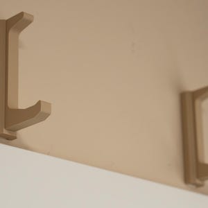 May include: Two beige wall-mounted hooks with a curved design. The hooks are mounted on a beige wall.