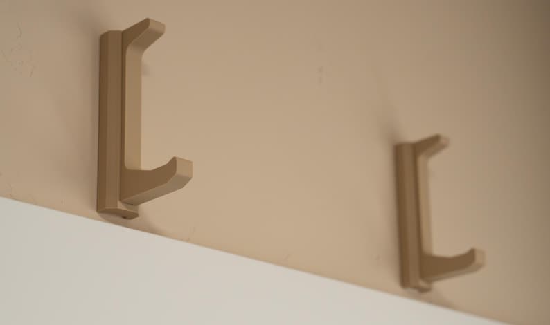 May include: Two beige wall-mounted hooks with a curved design. The hooks are mounted on a beige wall.