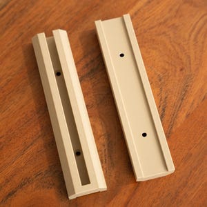 May include: Two beige plastic wall mount brackets with three holes each. The brackets are designed to hold a shelf or other object.