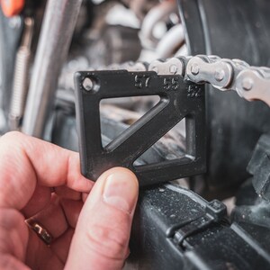 May include: A black plastic chain wear gauge with the numbers 52, 55, and 57 printed on it. The gauge is being used to measure the wear on a motorcycle chain.