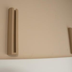 May include: Two beige wall mounted shelf brackets with a rectangular shape and a slot in the center.