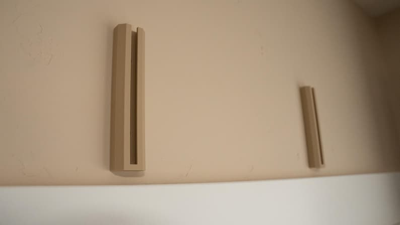 May include: Two beige wall mounted shelf brackets with a rectangular shape and a slot in the center.