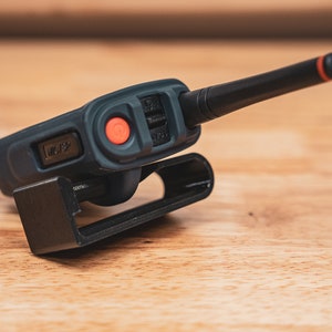 May include: A black walkie talkie with a red button and a black antenna. The walkie talkie is attached to a black holster.