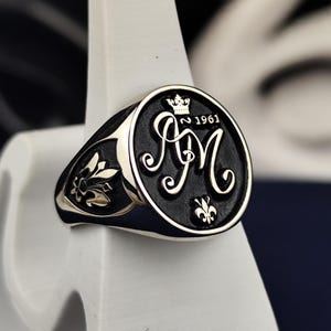 Könnte beinhalten: Ein silberner Siegelring mit einer runden schwarzen Fläche, die die eingravierten Buchstaben AM, eine kleine Krone und das Jahr 1961 zeigt. Die Seiten des Rings sind mit einem silbernen Fleur-de-Lis-Motiv auf schwarzem Grund verziert.