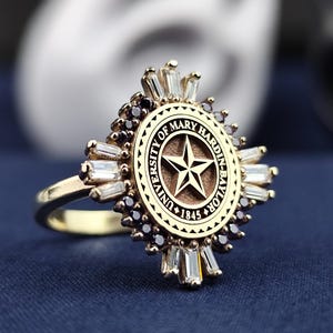 Op de afbeelding: Een gouden ring met een cirkelvormig ontwerp met het logo van de University of Mary Hardin-Baylor. Het logo is een ster met de woorden "University of Mary Hardin-Baylor" en "1845" eromheen. De ring is versierd met kleine zwarte en witte stenen.