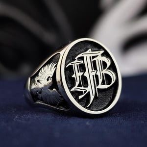 May include: A silver ring with a black enamel inlay featuring the initials "HFB" in a stylised font. The ring also has a detailed eagle design on the side.