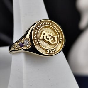May include: A gold class ring with a purple and white stone setting. The ring features the Abilene Christian University logo and the year 2025.