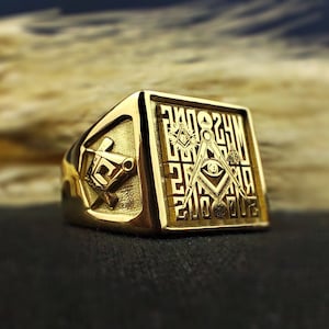 Puede incluir: Un anillo masónico de oro con un diseño cuadrado que presenta una brújula y una escuadra, un ojo que todo lo ve y otros símbolos masónicos.