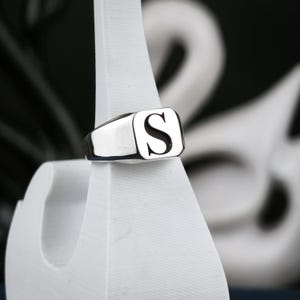 May include: A polished silver-tone signet ring featuring a flat, rectangular face engraved with a prominent, bold capital letter S in a serif font. The band is smooth and tapers slightly toward the back, showcasing a clean, metallic finish.