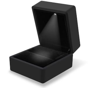 Könnte beinhalten: Eine schwarze Ringbox mit einem eingebauten LED-Licht, das das Innere beleuchtet. Die Box ist geöffnet und das Licht leuchtet auf den leeren Samt-Innenraum.