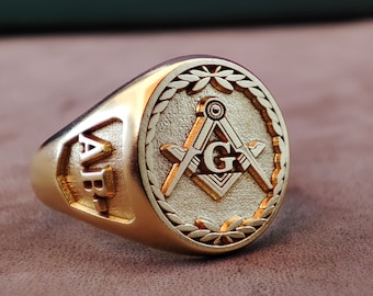 Può includere: Un anello color oro con una faccia circolare che presenta un simbolo massonico in rilievo composto da squadra e compasso con la lettera G al centro. Il motivo è bordato da una corona d'alloro. Il lato dell'anello mostra le lettere AB in rilievo.