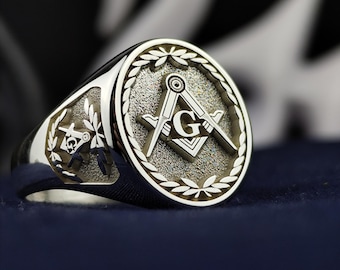 May include: A silver Masonic ring with a square and compass design and a letter G in the center. The ring is set with a laurel wreath around the design.