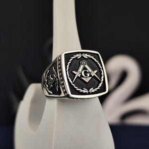 May include: A silver ring with a black square-shaped setting featuring a silver Masonic symbol with a 'G' in the center.
