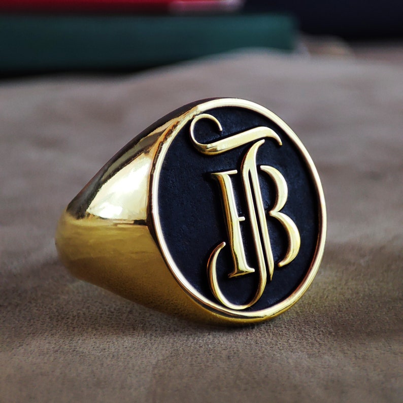 This detailed product image shows a personalized initial signet ring featuring a custom monogram design engraved on the flat top surface. The ring is offered in Gold Plated, 14k Gold, and Silver options, making it suitable for both men and women looking for a meaningful accessory. The engraving highlights the initials with clean, precise lines, emphasizing craftsmanship and timeless elegance. The photo clearly displays the polished finish, sturdy shape, and classic signet style.