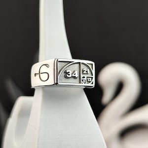 Puede incluir: Un anillo plateado con una cara rectangular que presenta la secuencia de Fibonacci. El anillo se muestra sobre un soporte blanco sobre un fondo oscuro. Los números 6, 34, 21, 13 y 8 son visibles en el anillo.