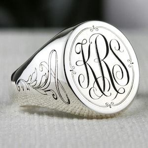 Può includere: Un anello sigillo in argento lucido con una faccia circolare che presenta un monogramma inciso con le lettere KRS in un carattere corsivo decorativo, circondato da un bordo sottile, con ulteriori incisioni a volute floreali sul lato.