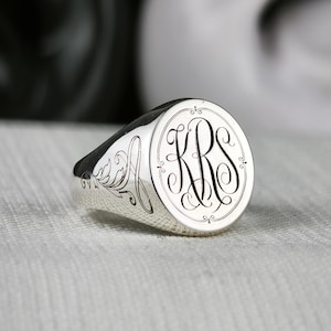 May include: A polished silver signet ring featuring a circular face engraved with the monogram letters KRS in a decorative script font. The side of the ring band displays an intricate, etched floral scrollwork pattern that extends from the face.