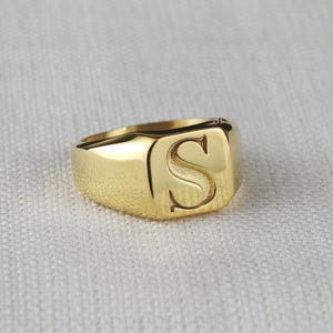 May include: A gold-toned signet ring featuring a polished finish and a prominent, recessed letter S engraved on the flat, rectangular top surface. The band is smooth and tapers slightly toward the back, showcasing a classic, solid metal construction.
