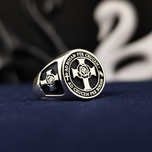 Puede incluir: Un anillo plateado con un diseño de cruz y rosa. El anillo presenta las palabras "AD CRUCEM PER ROSAM" alrededor de la cruz. El anillo está sobre una superficie azul oscuro.